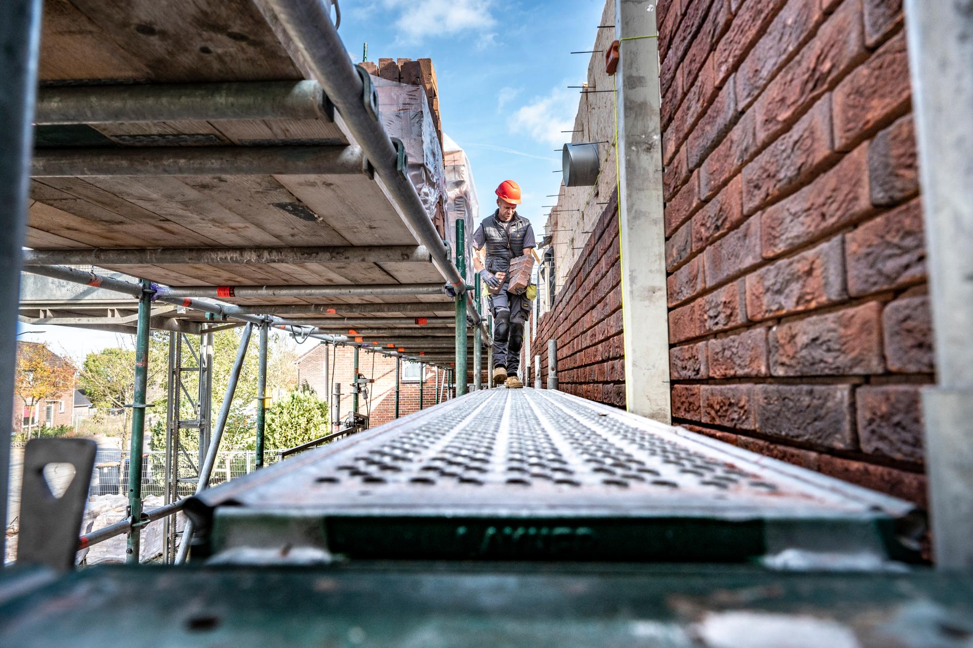 Man met rode helm die met bakstenen in z'n arme over een stijger loopt bij huizen in aanbouw