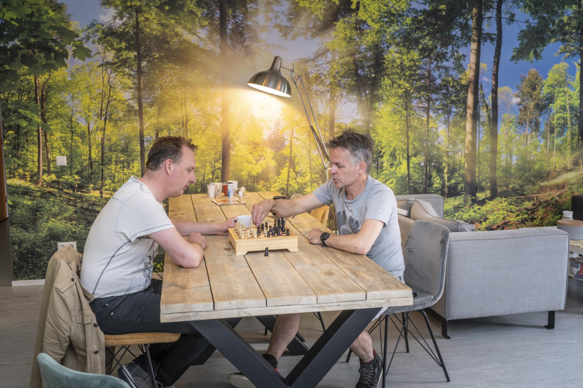 Tegen de achtergrond van een bosrijk behang zijn twee mannen aan het schaken aan een eettafel.